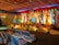 A beautifully decorated stage with vibrant floral arrangements and ornate backdrop. The stage is illuminated by colorful lights with red, yellow, and green hues. Chairs covered in white satin are arranged in rows facing the stage, and the ceiling is draped with multi-colored fabric, enhancing the festive atmosphere.