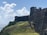 A historic stone fortress wall with a rugged, weathered appearance stands prominently on a grassy hillside. The fortress overlooks a body of water under a bright blue sky filled with fluffy white clouds. The texture of the stonework and the natural greenery around it add depth to the scene.