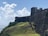 A historic stone fortress wall with a rugged, weathered appearance stands prominently on a grassy hillside. The fortress overlooks a body of water under a bright blue sky filled with fluffy white clouds. The texture of the stonework and the natural greenery around it add depth to the scene.