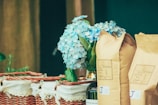 A wicker basket is adorned with a white cloth, containing several unopened brown paper bags, likely of coffee or similar product. A bouquet of light blue hydrangeas adds a touch of elegance and freshness to the arrangement.