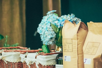 A wicker basket is adorned with a white cloth, containing several unopened brown paper bags, likely of coffee or similar product. A bouquet of light blue hydrangeas adds a touch of elegance and freshness to the arrangement.