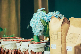 A wicker basket is adorned with a white cloth, containing several unopened brown paper bags, likely of coffee or similar product. A bouquet of light blue hydrangeas adds a touch of elegance and freshness to the arrangement.