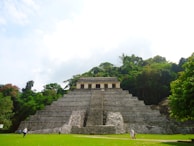 An ancient stone pyramid structure stands prominently in a lush jungle setting. The pyramid has a series of steep steps leading up to a small building or temple at the top. Greenery and dense trees surround the pyramid, creating a natural backdrop. Several people are visible on the ground level, walking around or standing near the pyramid.