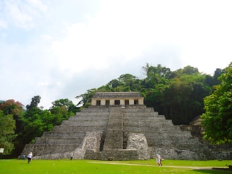 An ancient stone pyramid structure stands prominently in a lush jungle setting. The pyramid has a series of steep steps leading up to a small building or temple at the top. Greenery and dense trees surround the pyramid, creating a natural backdrop. Several people are visible on the ground level, walking around or standing near the pyramid.