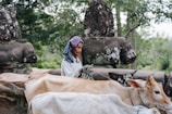 A person dressed in a traditional outfit leads two cows with ropes. In the background, there are ancient-looking stone statues surrounded by lush green trees.