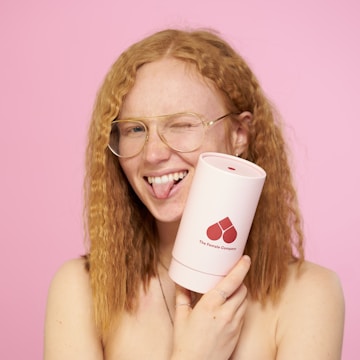A person with wavy red hair and wearing glasses is playfully sticking out their tongue, holding a pink cylindrical product with a red logo and the text 'The Female Company' against a pink background.