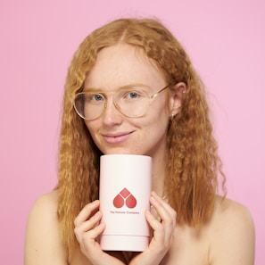 A person with curly red hair is holding a cylindrical container with the logo and text 'The Female Company' on it. The background is a plain, light pink color.