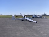 A sleek, white glider is positioned on an airstrip with its long wings extended. In the background, several other aircraft are parked near a grassy field. The sky is clear and blue, suggesting a calm weather setting.