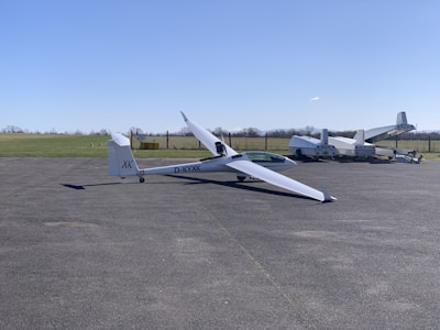 A sleek, white glider is positioned on an airstrip with its long wings extended. In the background, several other aircraft are parked near a grassy field. The sky is clear and blue, suggesting a calm weather setting.