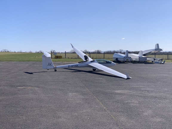 A sleek, white glider is positioned on an airstrip with its long wings extended. In the background, several other aircraft are parked near a grassy field. The sky is clear and blue, suggesting a calm weather setting.