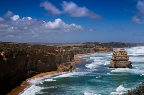 Cliffs formed from limestone stretch along a rugged coastline with a solitary rock stack rising from the ocean. Waves crash against the shore, with a sandy beach visible between the steep cliffs and the bright blue sea. Fluffy white clouds dot the sky, contrasting with the deep blue above.