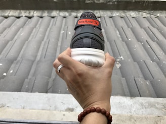 A hand holding a sneaker with a ribbed sole, a black and orange color scheme, and an orange tag reading 'Billy's Hunter X.' The hand has a ring on one finger and a beaded bracelet on the wrist. The background features a corrugated metal surface.