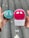 Two hands are holding skincare devices, one in each hand. The left hand holds a turquoise Foreo Luna Mini with ornate eye-like designs. The right hand holds a pink Foreo Luna Mini in its packaging with a similar design. The background suggests an outdoor environment with a blurred, neutral setting.