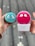 Two hands are holding skincare devices, one in each hand. The left hand holds a turquoise Foreo Luna Mini with ornate eye-like designs. The right hand holds a pink Foreo Luna Mini in its packaging with a similar design. The background suggests an outdoor environment with a blurred, neutral setting.