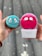 Two hands are holding skincare devices, one in each hand. The left hand holds a turquoise Foreo Luna Mini with ornate eye-like designs. The right hand holds a pink Foreo Luna Mini in its packaging with a similar design. The background suggests an outdoor environment with a blurred, neutral setting.