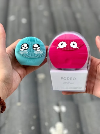 Two hands are holding skincare devices, one in each hand. The left hand holds a turquoise Foreo Luna Mini with ornate eye-like designs. The right hand holds a pink Foreo Luna Mini in its packaging with a similar design. The background suggests an outdoor environment with a blurred, neutral setting.