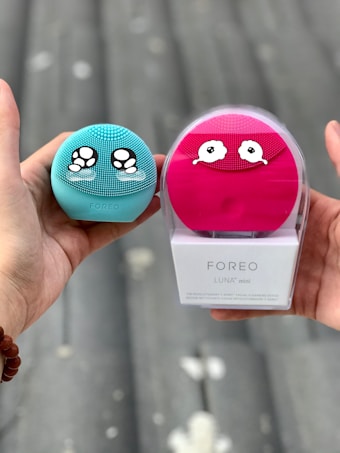 Two hands are holding skincare devices, one in each hand. The left hand holds a turquoise Foreo Luna Mini with ornate eye-like designs. The right hand holds a pink Foreo Luna Mini in its packaging with a similar design. The background suggests an outdoor environment with a blurred, neutral setting.