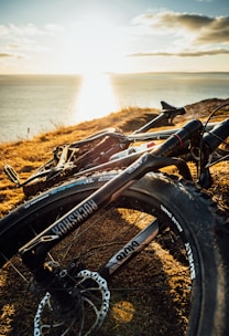 A mountain bike is situated on a grassy cliff overlooking a serene body of water. The sun is low on the horizon, casting a warm golden glow over the landscape and reflecting on the water. The bike's tires and frame are prominently displayed, showing details of the brand and components.