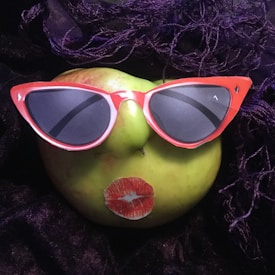 A green apple is humorously styled with cutouts of red sunglasses and lips, resembling a face. The apple is placed on a dark, textured fabric with purple strands enhancing the whimsical setting.