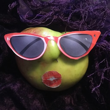 A green apple is humorously styled with cutouts of red sunglasses and lips, resembling a face. The apple is placed on a dark, textured fabric with purple strands enhancing the whimsical setting.