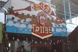 A decorative booth sign features an artistic representation of waves and abstract designs, with the word 'Tribe' prominently displayed in stylized letters. The sign incorporates vibrant colors and tribal patterns, complemented by a series of hanging bulbs beneath it. The background includes structural elements of the venue, such as a roof and walls, alongside some greenery.