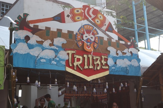 A decorative booth sign features an artistic representation of waves and abstract designs, with the word 'Tribe' prominently displayed in stylized letters. The sign incorporates vibrant colors and tribal patterns, complemented by a series of hanging bulbs beneath it. The background includes structural elements of the venue, such as a roof and walls, alongside some greenery.