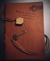 A brown leather notebook features an embossed design with scripted text and a postage stamp motif. It is wrapped with a leather cord that threads through the cover, adorned with a metallic key-shaped charm and a decorative plaque.
