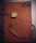 A brown leather notebook features an embossed design with scripted text and a postage stamp motif. It is wrapped with a leather cord that threads through the cover, adorned with a metallic key-shaped charm and a decorative plaque.