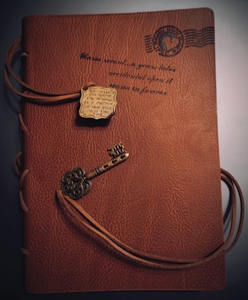 A brown leather notebook features an embossed design with scripted text and a postage stamp motif. It is wrapped with a leather cord that threads through the cover, adorned with a metallic key-shaped charm and a decorative plaque.