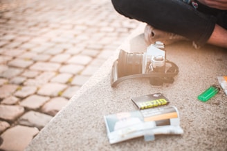 A vintage-style camera rests on a stone surface alongside a pack of film and a green lighter. The setting appears to be outdoors on a cobblestone street, with part of a person's legs visible, suggesting a casual or candid moment.