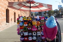 A street vendor stall displaying colorful knitted hats featuring a variety of character designs, positioned on a sidewalk near a brick building. A person dressed in a pink jacket and wearing a blue hat is standing nearby, facing the hats. The setting is urban with a partially visible street in the background.