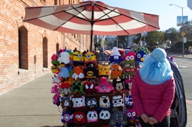 A street vendor stall displaying colorful knitted hats featuring a variety of character designs, positioned on a sidewalk near a brick building. A person dressed in a pink jacket and wearing a blue hat is standing nearby, facing the hats. The setting is urban with a partially visible street in the background.