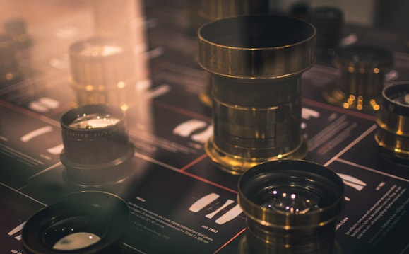 A display of vintage brass camera lenses is arranged on a reflective surface, surrounded by a softly blurred background of informational text and graphics, likely describing each lens.
