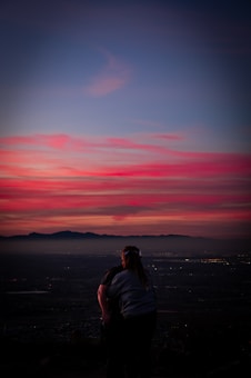 A couple stands closely together, facing a breathtaking sunset with vivid hues of pink, purple, and blue. In the background, a mountainous landscape under a darkening sky is visible, with city lights beginning to twinkle.