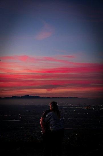 A couple stands closely together, facing a breathtaking sunset with vivid hues of pink, purple, and blue. In the background, a mountainous landscape under a darkening sky is visible, with city lights beginning to twinkle.