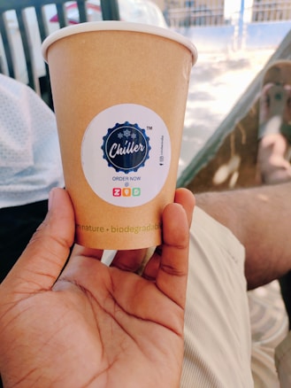 A hand holds a brown paper cup with a logo labeled 