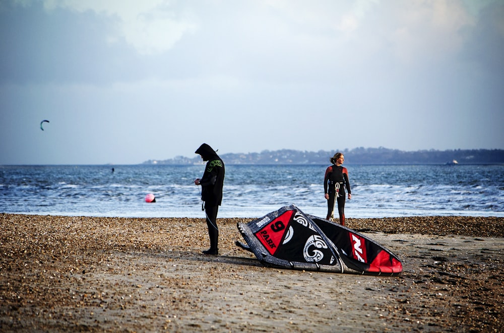 man in black jacket standing beside red and black kayak on beach during daytime
