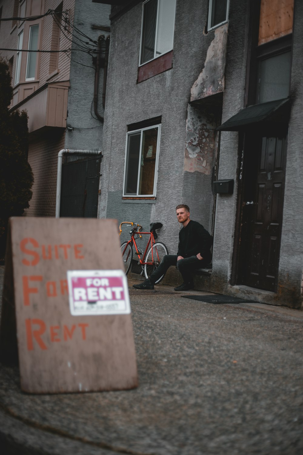 man in black jacket and blue denim jeans sitting on black wheelchair beside brown concrete building