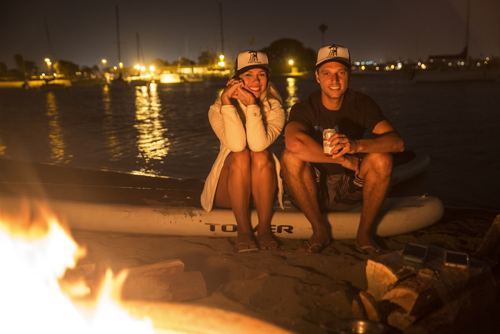 Hombre y mujer sentados en arena marrón cerca del cuerpo de agua durante la noche