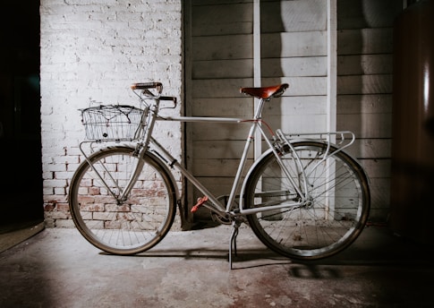 A vintage-style bicycle with a basket is parked against a shabby chic wooden wall. The bicycle features a leather saddle and handlebars, with its frame painted in a light cream color. The background consists of white painted brick and wooden boards, adding a rustic charm to the setting. Subtle shadows create depth and highlight the texture of both the wall and the bicycle.