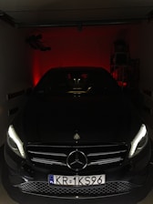 A sleek, black Mercedes-Benz car is parked in a dimly lit garage. The headlights are on, casting a soft glow and highlighting the car's front grill and emblem. The background is dark, with a faint red light creating a dramatic effect.