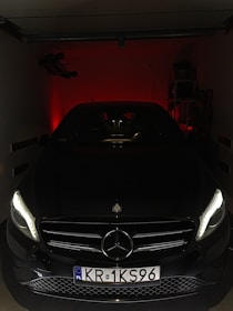A sleek, black Mercedes-Benz car is parked in a dimly lit garage. The headlights are on, casting a soft glow and highlighting the car's front grill and emblem. The background is dark, with a faint red light creating a dramatic effect.