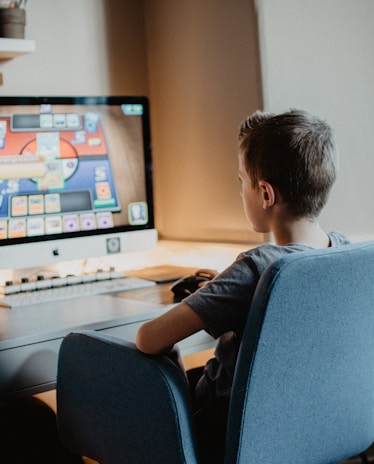 A young person is sitting on a blue chair at a desk, focused on a computer screen displaying a colorful card game. The scene appears to be set in a cozy, well-lit room.