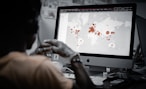 A person is viewing a map with red data points on a computer monitor, likely indicating a geographical distribution. The image has a focus on technology and data analysis.