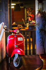 A bright red vintage scooter is parked in an indoor setting, leaning against a white wall. The scooter has a visible license plate. Behind it is a bar area with stools and tables, and some plants on the right side. The indoor lighting creates a warm and cozy environment.