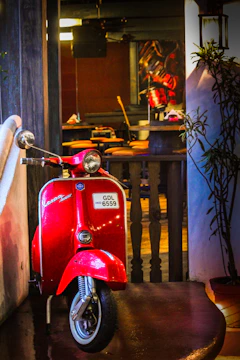 A bright red vintage scooter is parked in an indoor setting, leaning against a white wall. The scooter has a visible license plate. Behind it is a bar area with stools and tables, and some plants on the right side. The indoor lighting creates a warm and cozy environment.