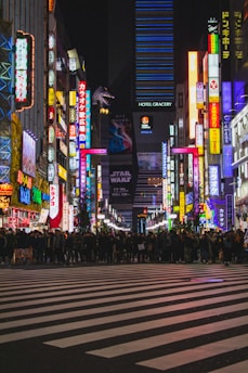 A bustling city scene with neon lights illuminating a crowded street. Pedestrians gather at a zebra crossing in an urban nightlife area. Bright colorful signs and billboards line the street, depicting various advertisements. A large Godzilla figure can be seen atop one of the buildings, with 'Hotel Gracery' prominently displayed. Movie advertisements, including Star Wars, are visible.