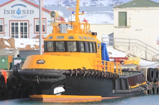 A large, bright yellow and black tugboat docked at a snowy harbor. The harbor features an industrial building in the background with the words 'ISTUSID' visible. The boat has a sturdy rubber bumper along its sides and various equipment on deck. Snow covers parts of the surrounding area.