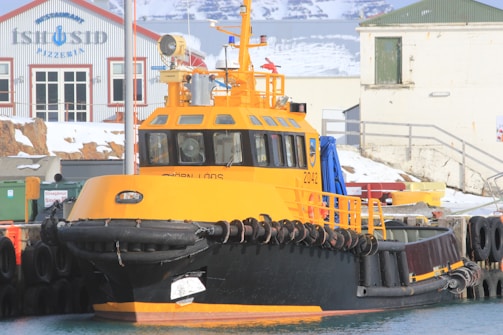 A large, bright yellow and black tugboat docked at a snowy harbor. The harbor features an industrial building in the background with the words 'ISTUSID' visible. The boat has a sturdy rubber bumper along its sides and various equipment on deck. Snow covers parts of the surrounding area.
