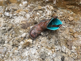 A single brown cowboy boot with turquoise interior lies on rocky and uneven ground. There are scattered rocks and debris surrounding the boot, giving an impression of a rugged or outdoor setting. A couple of dry leaves are also visible among the rocks.